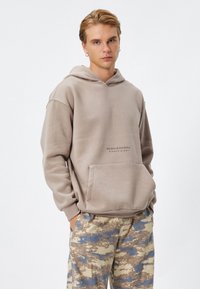 Sudadera con capucha beige y bolsillo delantero, con el texto "CREE EN TI MISMO" en una impresión oscura, combinada con pantalones cargo de estampado.