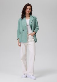 Mintgrønn blazer med doble knapper og klaffelapper, sammen med hvite vide bukser og hvite sneakers på en nøytral bakgrunn.