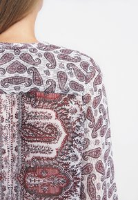 Transparente Bluse mit einem filigranen roten Paisleymuster auf einem weißen Hintergrund, mit leichter Textur und entspannter Passform.
