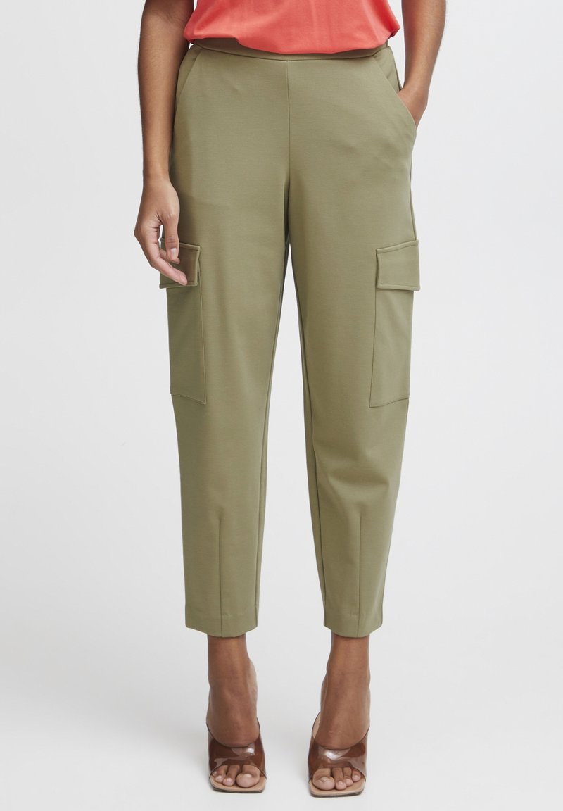 Femme portant un pantalon cargo court vert olive avec des poches latérales, associé à des talons ouverts marron et un haut corail, debout devant un fond uni.