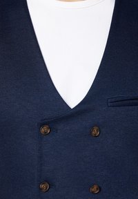 Granatowa kamizelka dwurzędowa z teksturowanej tkaniny, z brązowymi guzikami i małą kieszonką po lewej stronie, noszona na białej koszuli.