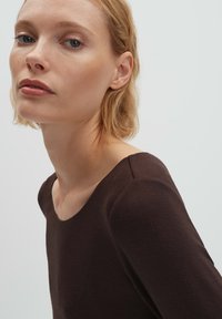 Brauner Langarmpullover mit einem runden Ausschnitt, weichem Material, figurbetontem Design und Nahtdetails an den Schultern. Einheitlicher Hintergrund.