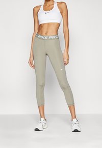 Nike Pro cropped leggings i olivengrøn med elastisk talje, der har lysegrå "NIKE PRO" tekst, parret med en hvid sports-bh.