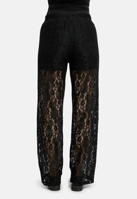 Juodos nėrinės kelnės turi pusiau permatomą dizainą su gėlių raštu, plataus kojų silueto ir elastingą liemens juostą. Nugaros vaizdas pabrėžia sudėtingas detales.