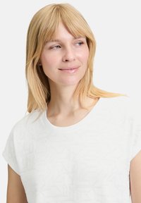 Hvid kortærmet top med et geometrisk præg i stoffet, rund halsudskæring og blødt bomuldsmateriale. Den subtile tekstur tilføjer visuel interesse.