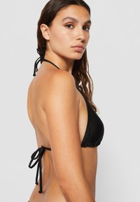 Čierny bikiny top s trojuholníkovým dizajnom, nastaviteľným zaväzovaním za krkom a na chrbte, vyrobený z hladkej látky, predvádzajúci jednoduchý, minimalistický štýl.