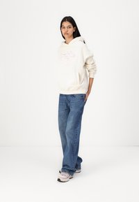 Beige hoodie with a graphic design, front pocket, paired with blue straight-leg jeans and light pink sneakers with black soles.