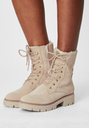 Botas de tobillo de ante beige con forro de pelo, ojales dorados y suela resistente. Presentan cordones y una cremallera lateral para facilitar su uso.