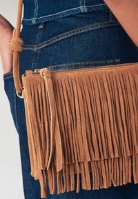 Borsa a bandoliera in suede beige con dettagli in frange verticali lunghe. Chiusura con zipper e tracolla beige, appoggiata su un tessuto di denim blu.