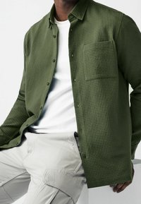 Groen gemêleerd opgestikt overhemd met een zak en een witte crew-neck t-shirt eronder, gecombineerd met lichtgrijze cargobroek.