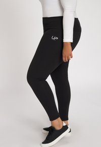 Zwarte leggings van rekbare stof met een hoge tailleband en een wit "UP Sports" logo op de heup, gecombineerd met zwarte sneakers.