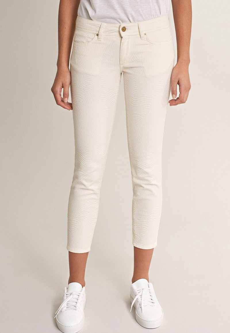 Femme portant un pantalon slim raccourci de couleur blanc cassé, des baskets blanches et un t-shirt blanc rentré, debout devant un fond uni.
