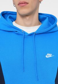 Modra Nike kapuca s kontrastnimi temnimi detajli, ki vključuje kapuco na vrvico, teksturirano tkanino in majhen bel logotip na prsih.