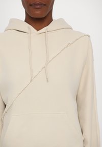 Beige hoodie with a raw edge diagonal seam, drawstrings, and a front pocket. Made of soft, textured fabric.