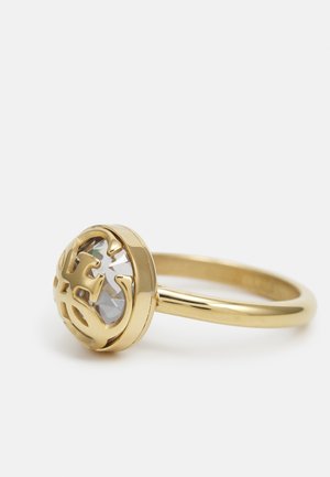 Gouden ring met een ronde heldere geslepen steen, gedeeltelijk bedekt door een decoratieve gouden logo-overlay op een gladde band.