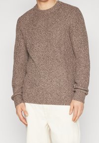 Sweter wełniany w ciepłym brązowym kolorze z fakturowanym wzorem, okrągłym dekoltem i ściągaczami na rękawach, połączony z jasnymi spodniami.