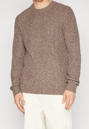 Suéter de punto en un cálido color marrón con un patrón texturizado, cuello redondo y puños de canalé, combinado con pantalones de color claro.