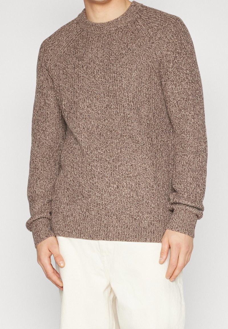 Sweter wełniany w ciepłym brązowym kolorze z fakturowanym wzorem, okrągłym dekoltem i ściągaczami na rękawach, połączony z jasnymi spodniami.