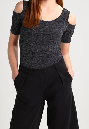 Femme aux cheveux roux portant un haut épaules dénudées gris foncé scintillant et un pantalon noir taille haute à jambes larges, les mains dans les poches.