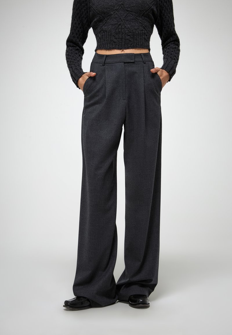 Hoge taille, donkergrijze broeken met een op maat gemaakte pasvorm, plooien en zijzakken. De stof heeft een gladde textuur, gecombineerd met glanzende schoenen.