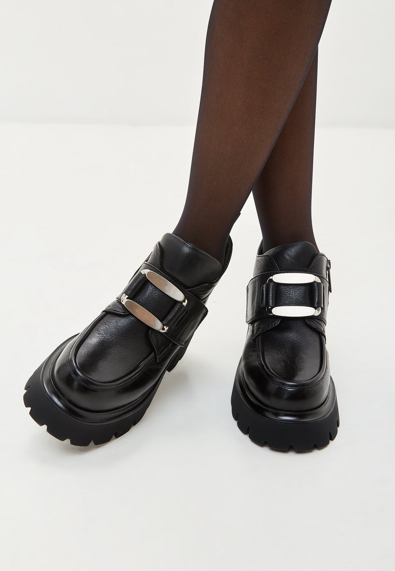 Zapatos de cuero negro con suela gruesa, con dos hebillas metálicas y una textura suave. El diseño incluye una puntera redondeada y un tobillo alto.