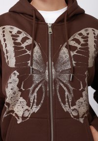 Brūns rāvējslēdzēja džemperis ar kapuci, uz kura ir liela, mirdzoša tauriņa grafika. Materiāls ir mīksts, ar aizskrūvējamiem auklām un metāla rāvējslēdzēja detaļām.