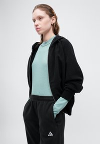 Μαύρη φούτερ ζακέτα με άνετη εφαρμογή, στρωμένη πάνω από ένα ανοιχτό μπλε ρι�γέ μπλούζα. Μαύρο παντελόνι φόρμας με τριγωνικό λογότυπο.