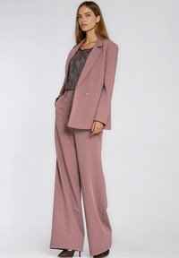 Kobieta w różowofioletowym garniturze z szerokimi spodniami i dwurzędową marynarką oraz brązowymi butami na wysokim obcasie, pod marynarką czarna bluzka w wzory.