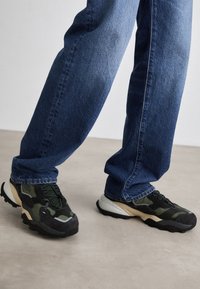 Grønne og svarte sneakers med teksturerte overlegg og en kraftig såle, kombinert med mørkeblå rette bukser, mot en nøytral bakgrunn.