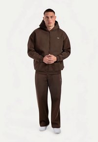 Brązowa bluza z kapturem na zamek i pasujące spodnie wykonane z miękkiego materiału, z małym logo na piersi oraz białe trampki.