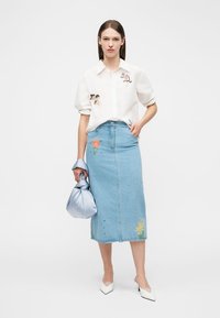 Kvinde iført hvid skjorte med broderede motiver, lyseblå denimnederdel med blomsterbroderi, hvide hæle og holder en lyseblå satin taske.