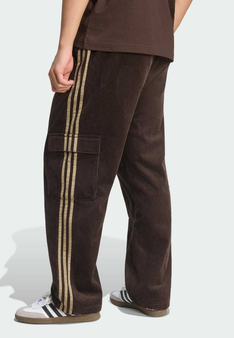Person iført brune fløjlsbukser med beige sidelister og cargolomme, kombineret med hvide sneakers med sorte striber.
