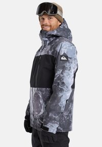 Mann in grauer und schwarzer gemusterter Winterjacke, beiger Strickmütze, schwarzen Skihandschuhen und schwarzen Skibrillen auf der Stirn, der seitlich lächelt.