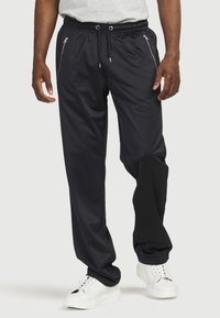 Schwarze Sporthose mit lockerer Passform, ausgestattet mit einem Kordelzug am Bund und Reißverschlusstaschen an den Seiten, aus glattem, leichtem Material.