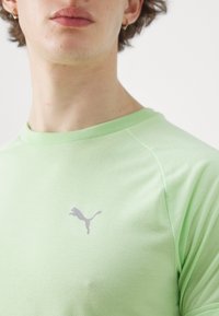 Camiseta verde claro con cuello redondo y mangas raglán cortas. Presenta un pequeño logo gris de Puma en el pecho. Textura de tela suave.