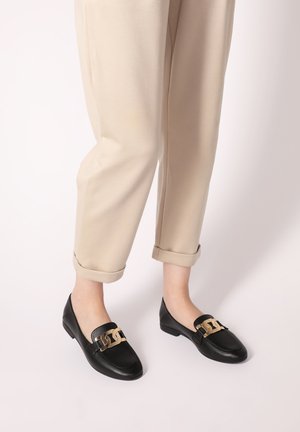 Schwarze Lederloafers mit einem goldenen Akzent am Riemen, featuring einer abgerundeten Zehenpartie und einem niedrigen Blockabsatz, getragen mit hochgerollten beigen Hosen.