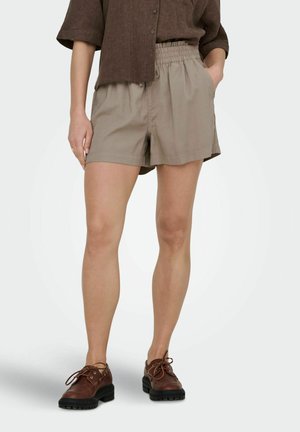 Kobieta w beżowych szortach z elastycznym pasem, brązowej koszuli na guziki i brązowych sznurowanych butach, stojąca z jedną ręką w kieszeni.