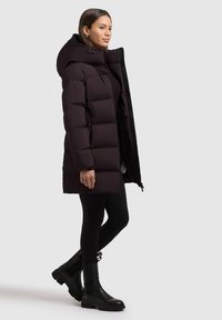 Abrigo de plumas morado oscuro con capucha, que presenta un cierre de cremallera en el frente y detalles elásticos, combinado con leggings negros y botas de tobillo negras resistentes.