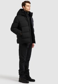 Chaqueta acolchada negra con capucha, con cierre de cremallera en la parte delantera, puños acanalados y dos bolsillos laterales, combinada con jeans oscuros y zapatos negros.