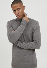 Grauer Rollkragenpullover, gerippte Textur, taillierter Schnitt, lange Ärmel, dezente Saumdetails, getragen mit verschränkten Armen vor einem hellen Hintergrund.