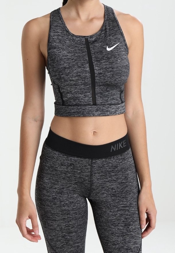 Femme portant un soutien-gorge de sport Nike gris foncé et des leggings taille haute assortis, debout devant un fond clair uni.