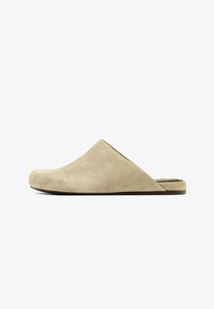 Beżowy zamszowy klapek wsuwany typu mule z zamkniętym, zaokrąglonym noskiem i czarną, płaską podeszwą, widoczny z boku na białym tle.