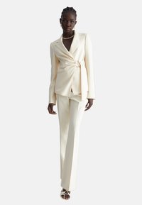 Completo su misura color crema con cintura in vita, maniche lunghe e pantaloni a gamba dritta. Tessuto liscio con dettagli minimi.