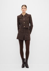 Giacca corta a quadri marrone con bottoni dorati abbinata a una gonna a quadri coordinata. Modella indossa collant marroni e stivaletti alla caviglia.
