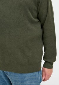 Pull en olive texturé avec des manches longues et une finition côtelée. Associé à un jean bleu clair, vu de côté avec l'étiquette de la marque.