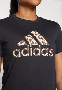 Svart bomullst-shirt med en stor beige leopardmönstrad Adidas-logga på framsidan. Korta ärmar och rund halsringning. Mjuk textur.