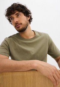 Uomo con capelli ricci e barba che indossa una maglietta verde oliva, appoggia gli avambracci su una superficie di legno, con uno sguardo contemplativo su uno sfondo semplice.