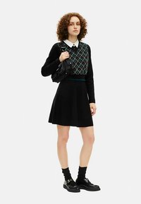 Schwarzer Rock kombiniert mit einem schwarzen und türkisfarbenen Argyle-Muster-Pullover über einem weißen Hemd mit Kragen. Komplettiert mit schwarzen Schuhen und einer kleinen Tasche.