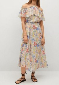 Robe maxi florale avec décolleté à volants sur les épaules, présentant une taille élastique, un tissu léger, et un mélange de couleurs pastel et de motifs.