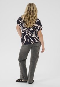Femme avec de longs cheveux blonds ondulés portant un top à motifs noir et beige, un jean gris et des sandales noires, se tenant avec une main sur la hanche.